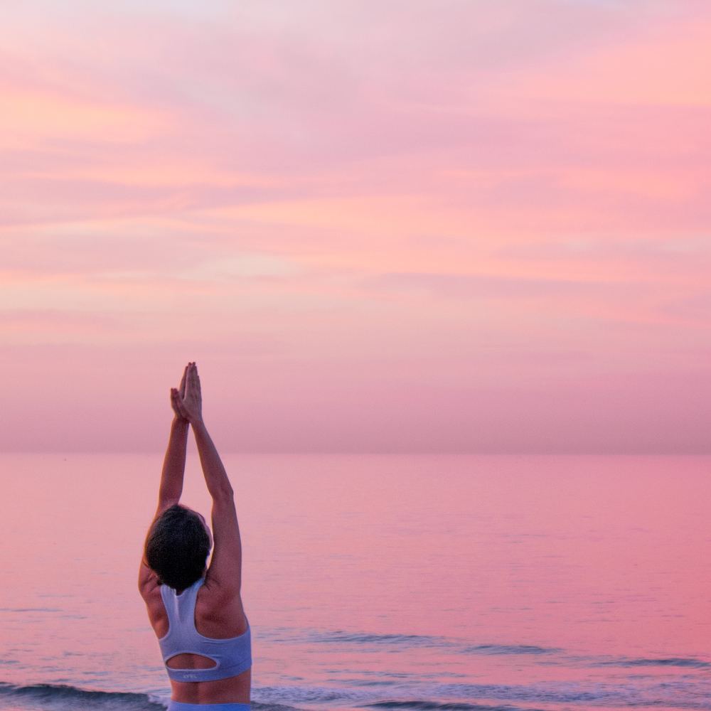 Issy Luck standing with arms overhead in a sunrise yoga practice beside the sea